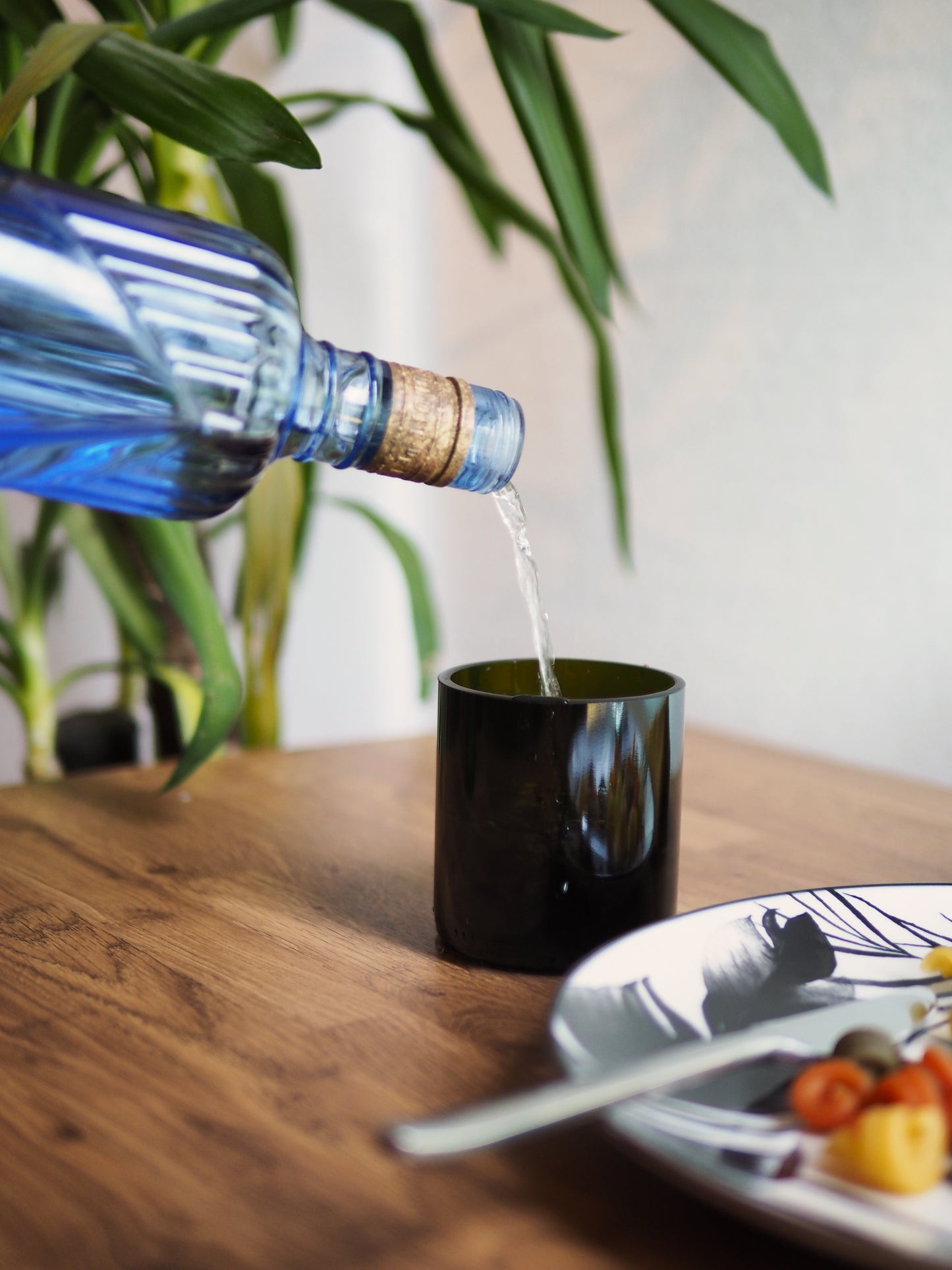 Verre vert foncé fabriqué à partir d'une bouteille recyclée, posé sur une table en bois. Eau versée depuis une bouteille en en verre bleu, scène de table chaleureuse avec plat de pâtes et décoration végétale. Accessoire écoresponsable pour une table durable et design.