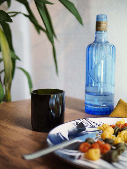 Verre vert foncé fabriqué à partir d'une bouteille recyclée, posé sur une table en bois. Scène de table chaleureuse avec plat de pâtes et décoration végétale. Accessoire écoresponsable pour une table durable et design.