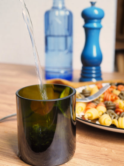 Verre vert foncé fabriqué à partir d'une bouteille recyclée, posé sur une table en bois. Eau versée, scène de table chaleureuse avec plat de pâtes. Accessoire écoresponsable pour une table durable et design.